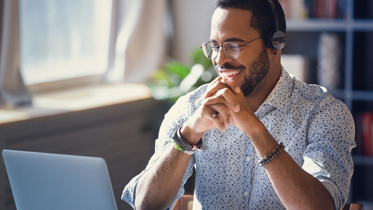 A man with headphones sits at a desk, focused on his laptop, engaged in a virtual meeting or task.