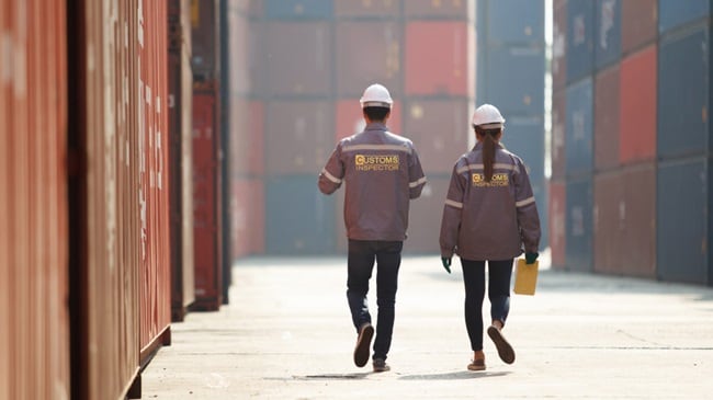 Two customs inspectors wearing helmets and uniforms walk through a shipping container yard.