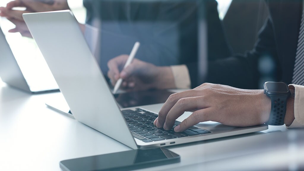 A person in a suit is focused on typing on a laptop at a desk, surrounded by office items.
