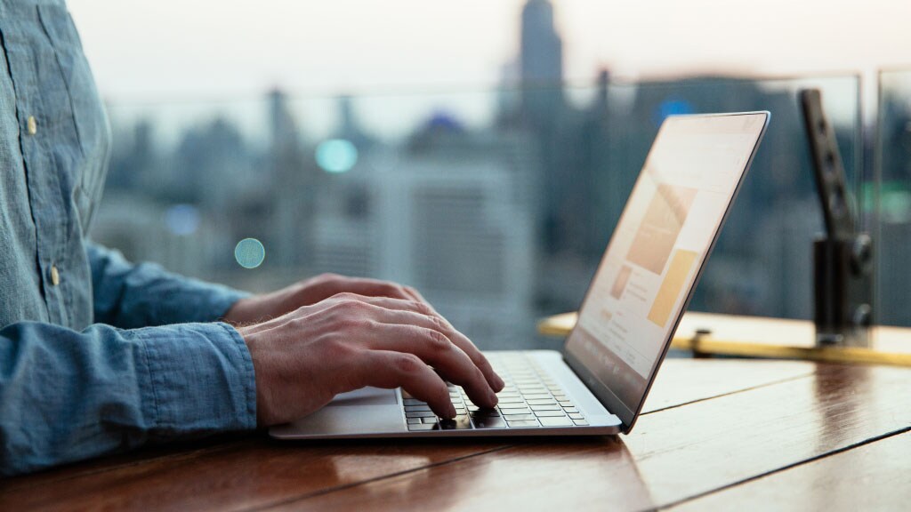 Individual focused on a laptop, with a vibrant cityscape behind them, blending work and urban scenery.