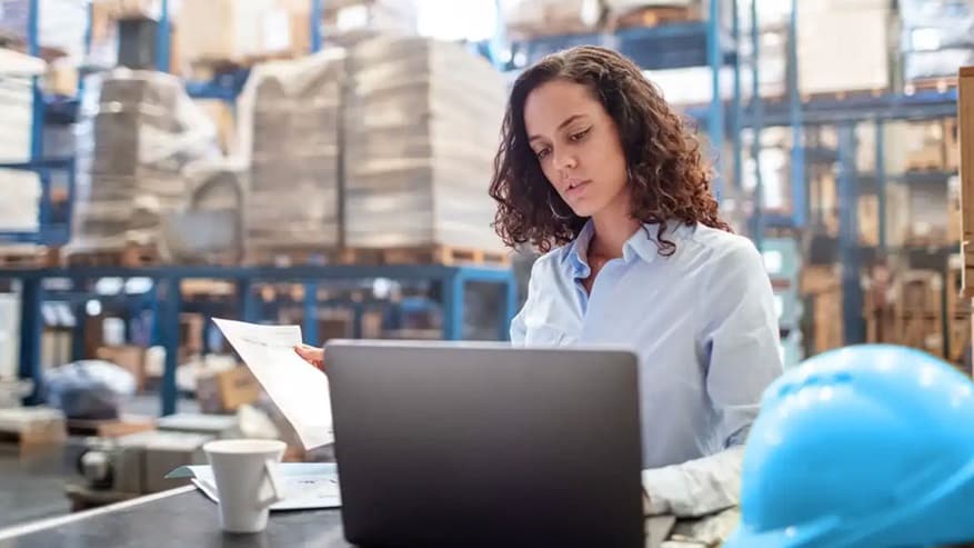 A woman sitting in a warehouse holding a document and looking at a laptop.