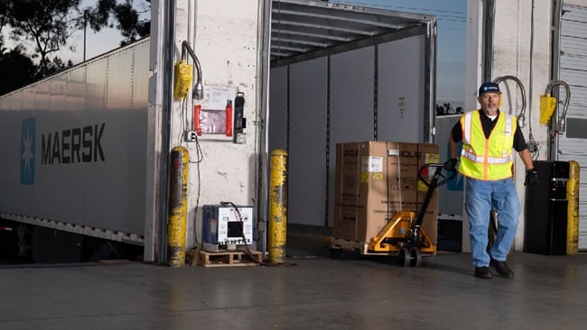 Person in a high-visibility vest using a manual pallet jack to move a pallet loaded with boxes