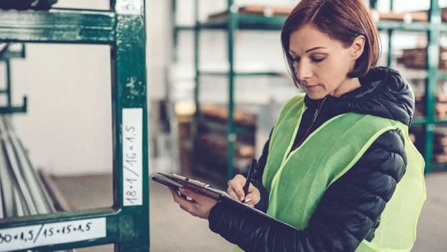 A woman with short hair wearing a black jacket and green safety vest in a warehouse reviewing papers on a writing pad.