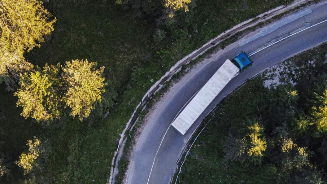 Vista aérea de um caminhão percorrendo uma estrada sinuosa rodeada por campos abertos verdejantes.
