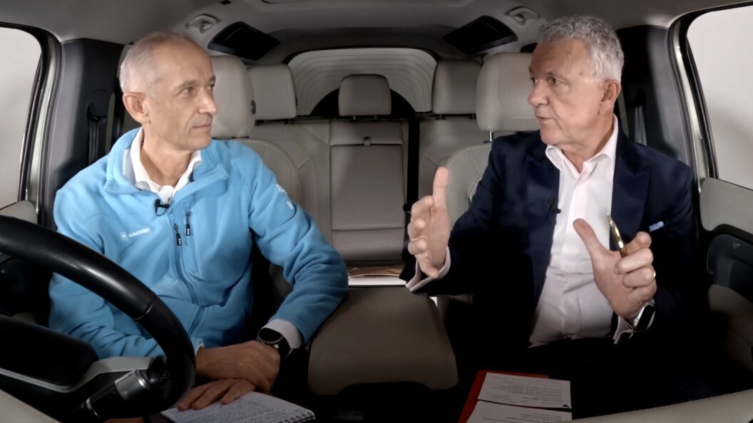 Two men sitting in the driver's seat of a car, both focused on the road ahead.