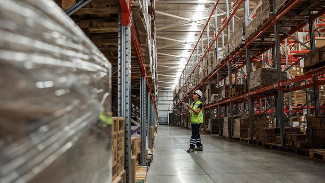 A worker stands in the center of a warehouse aisle, with shelves stocked on either side.