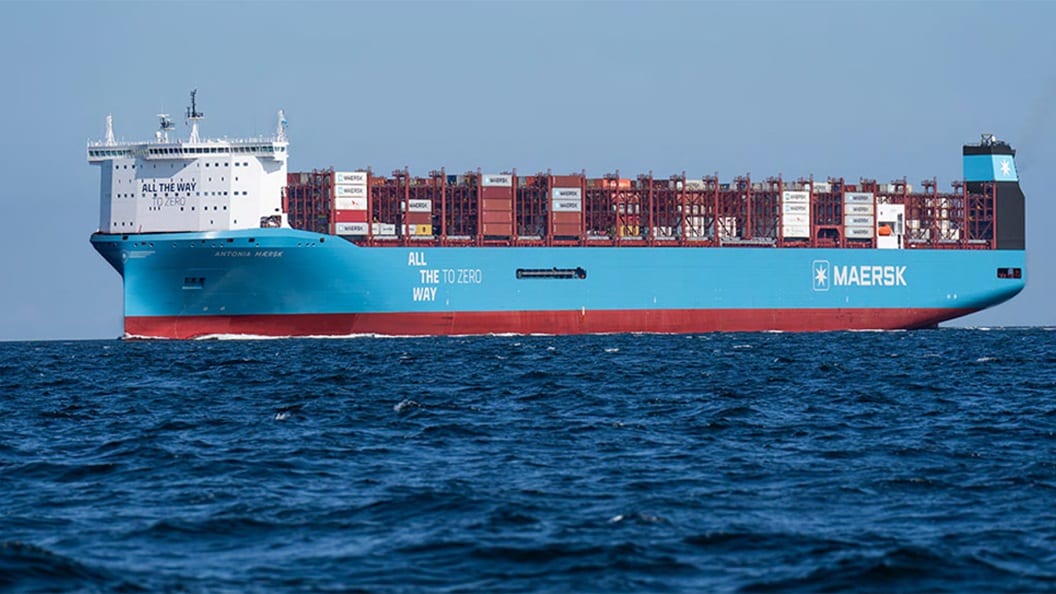 A large Maersk container ship, painted blue and red and loaded with shipping containers, sails on the open sea under clear skies.
