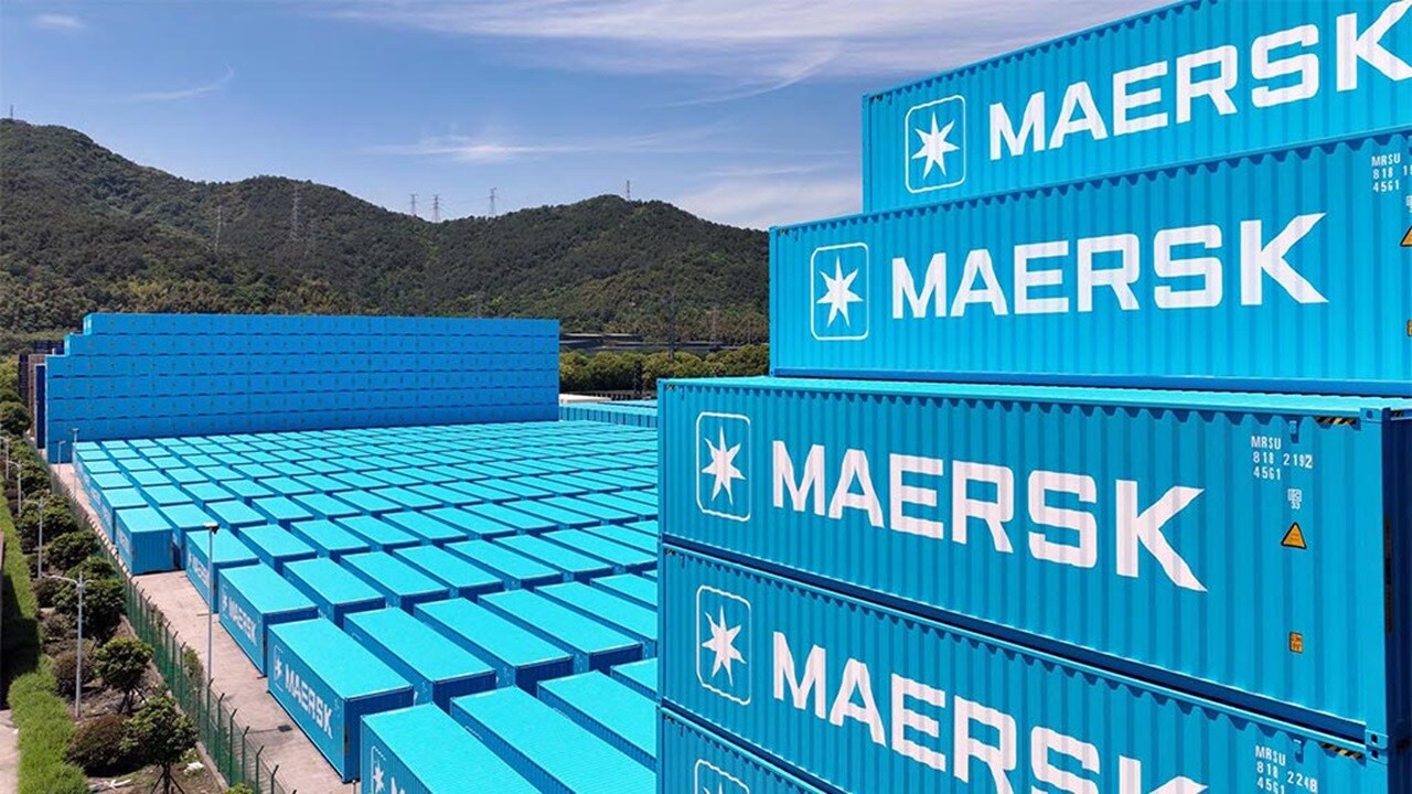 Stacks of blue Maersk shipping containers are arranged in rows at a storage facility, with green hills and a partly cloudy sky in the background.