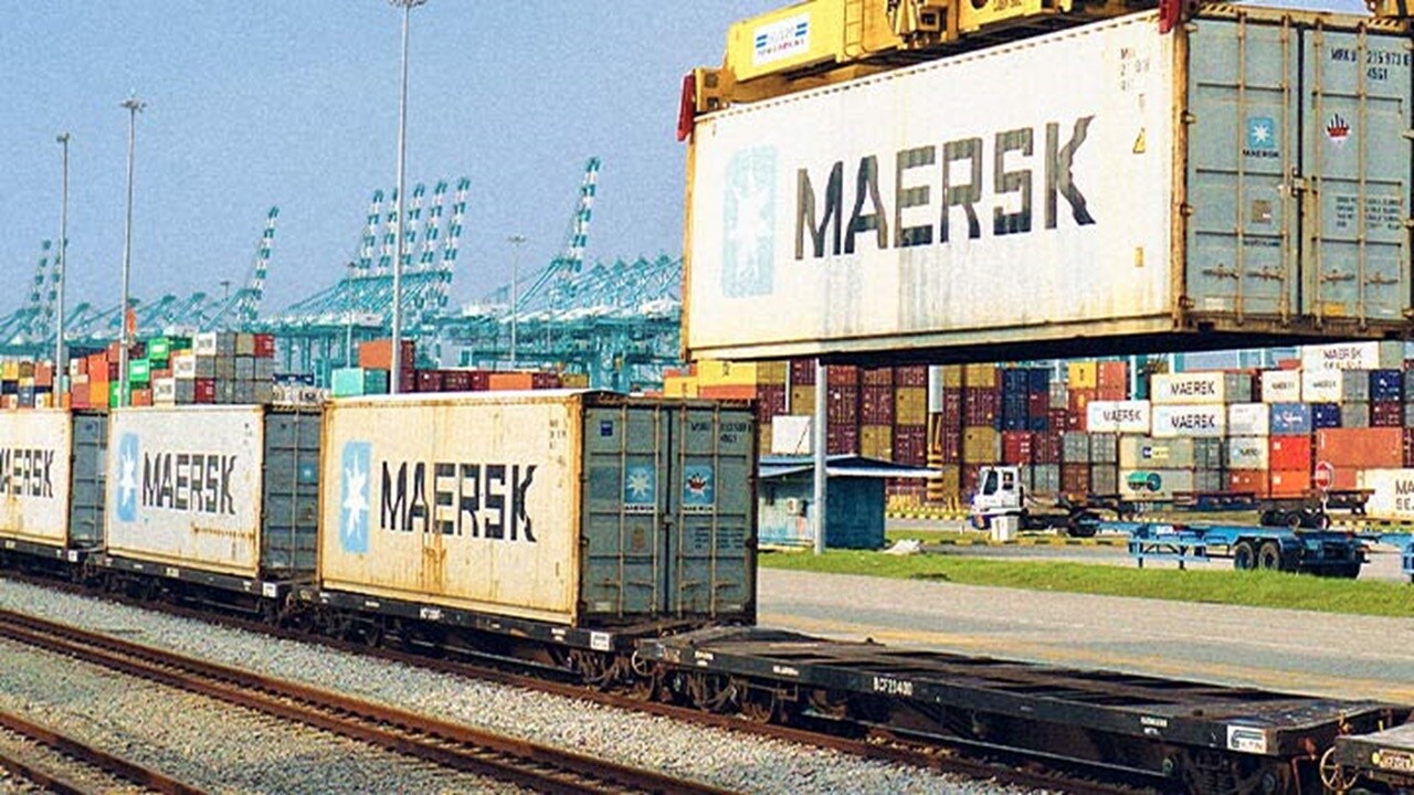 Maersk containers being loaded onto a block train at a rail terminal, with cranes and stacked containers in the background.