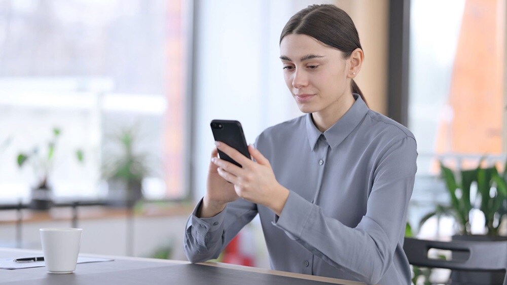 Woman in a modern office using a smartphone while sitting at a desk with a coffee cup, focused on her screen.