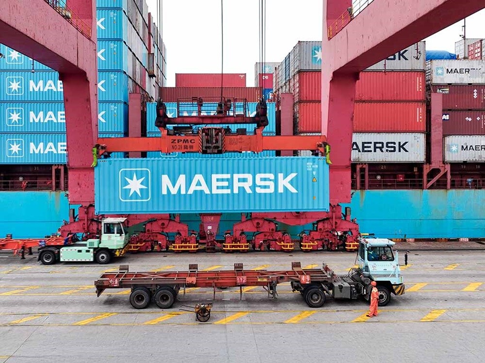 Un container Maersk in fase di sollevamento tramite gru in un terminal portuale operativo, con camion in transito nell'area sottostante