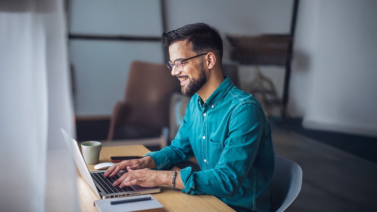 Smiling professional wearing glasses working on a laptop at a desk with a coffee mug nearby