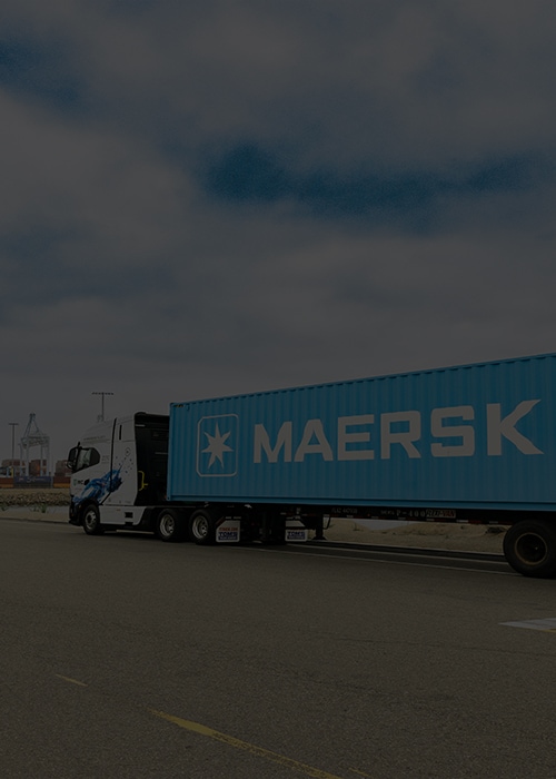 A blue Maersk truck is parked on a road near an industrial port, with shipping containers and cranes visible in the background under a cloudy sky.
